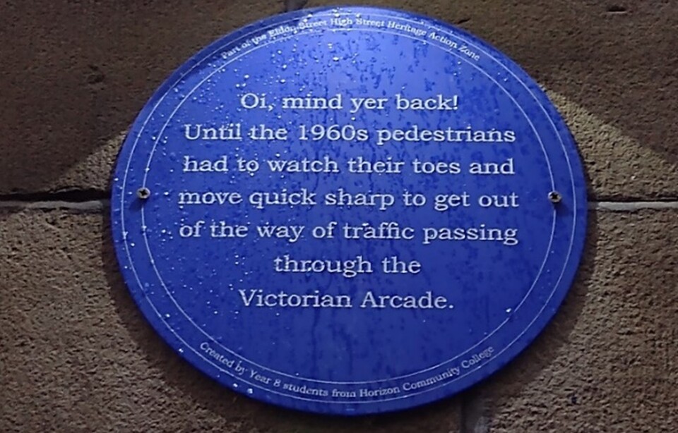 A blue circular plaque on a stone wall reads: "Oi, mind yer back! Until the 1960s pedestrians had to watch their toes and move quick sharp to get out of the way of traffic passing through the Victorian Arcade.